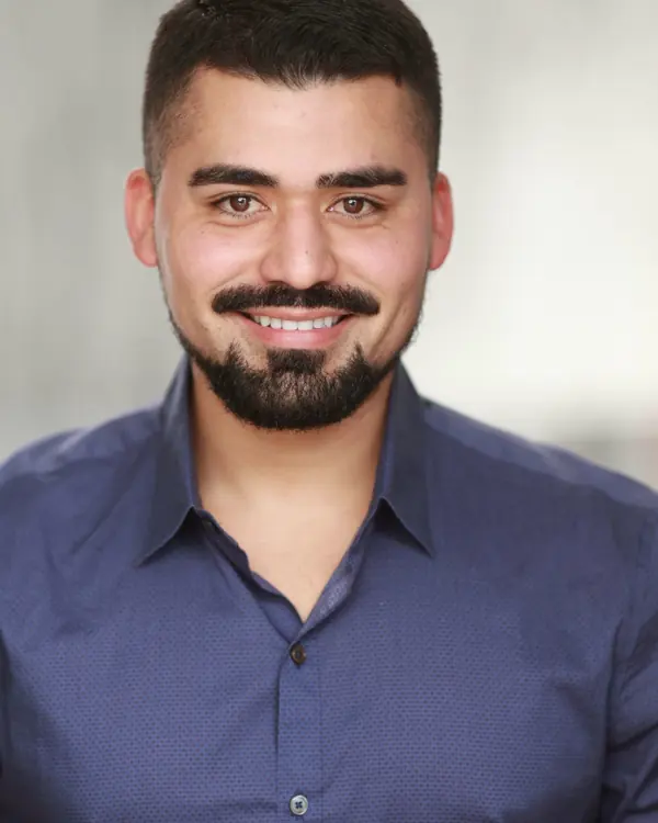 A smiling man with short dark hair and a neat beard in a blue button-down shirt, against a pale grey backdrop.