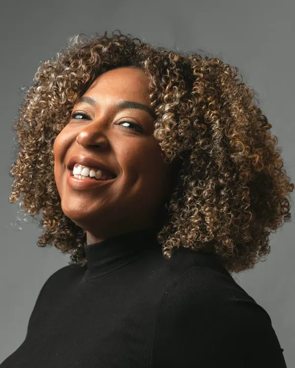 A smiling woman with shoulder-length curly hair in a black top, against a soft grey wall.