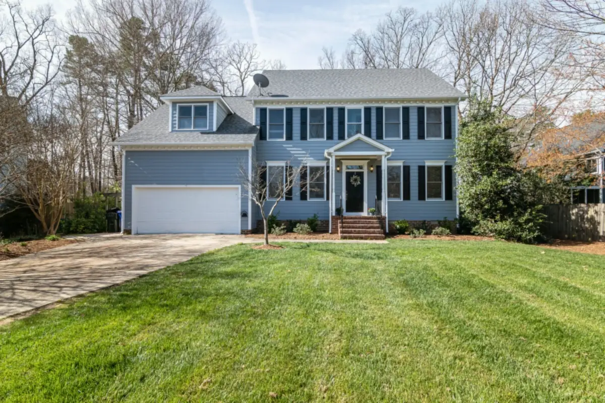 A blue two-story colonial with black shutters, a small front portico and an attached two-car garage, behind a freshly mown lawn in early spring.