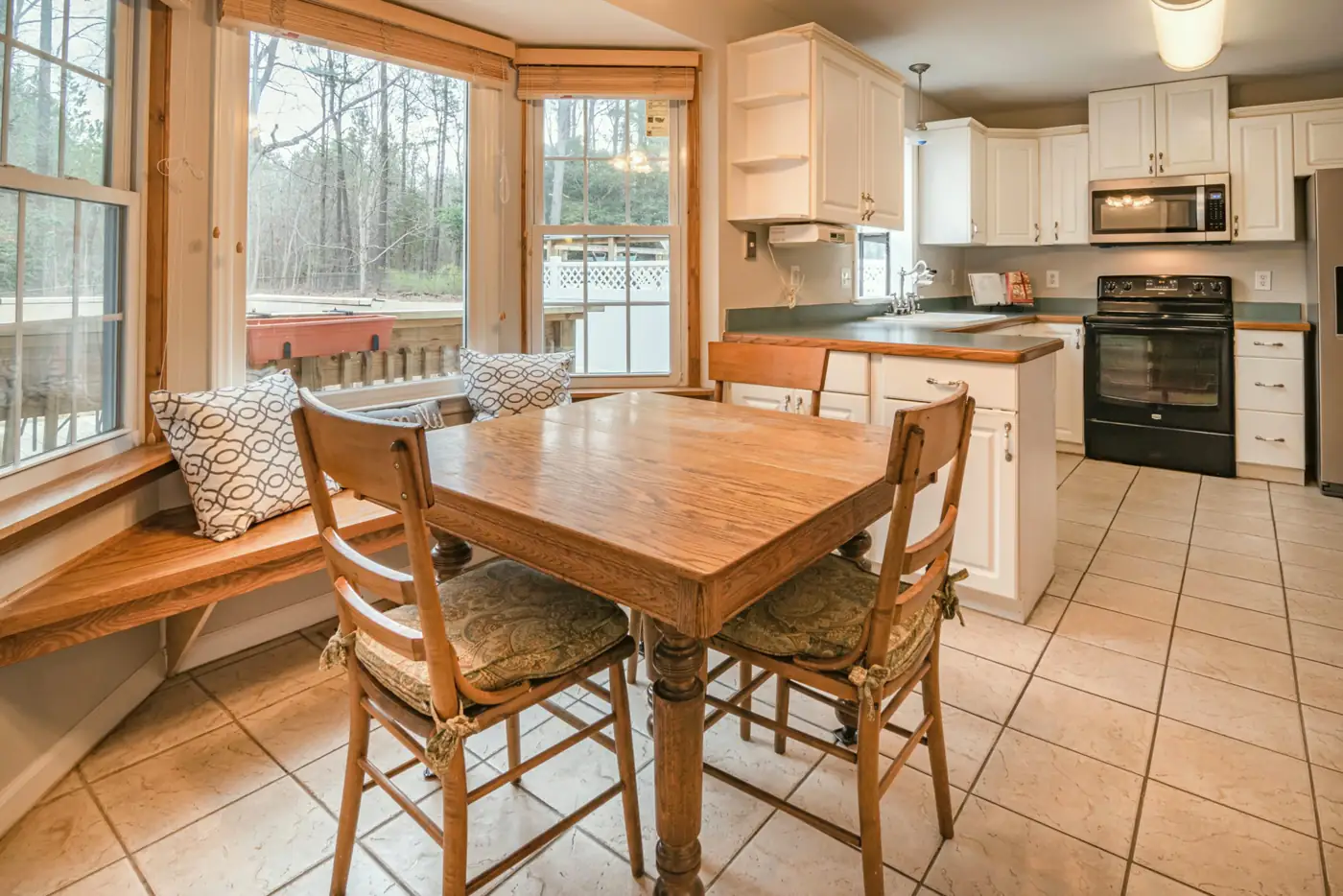 A breakfast nook with a built-in window bench and an oak table beside a white kitchen, looking out to the back deck.