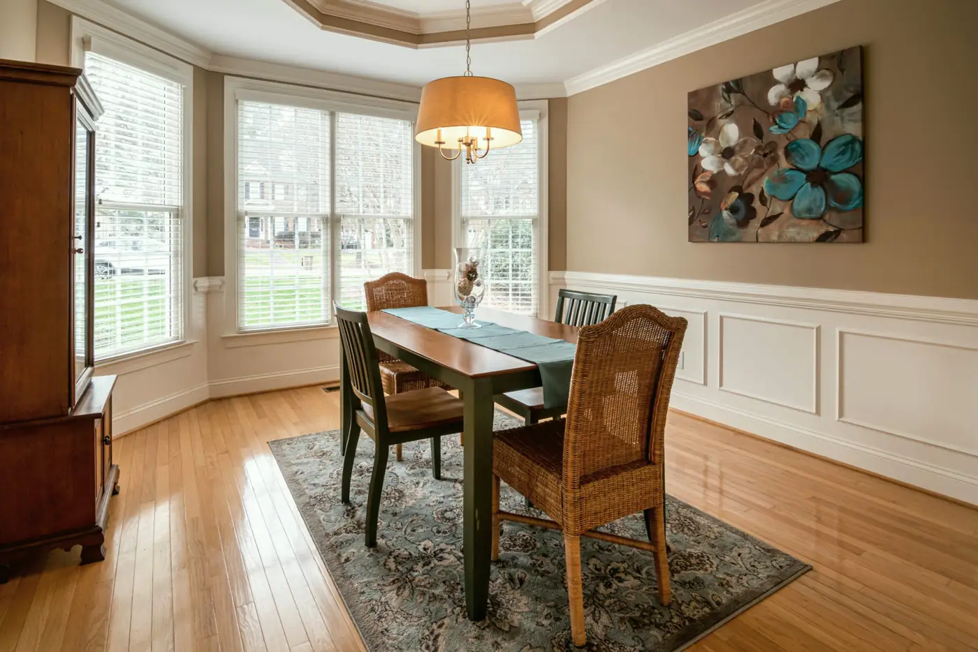 A dining room with a bay of three windows, white wainscoting, a tray ceiling and a table for four on a patterned rug.