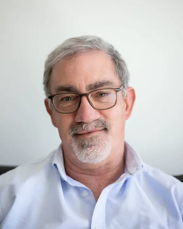 A smiling man in his sixties with grey hair, a trimmed beard and glasses, in a light blue open-collar shirt.