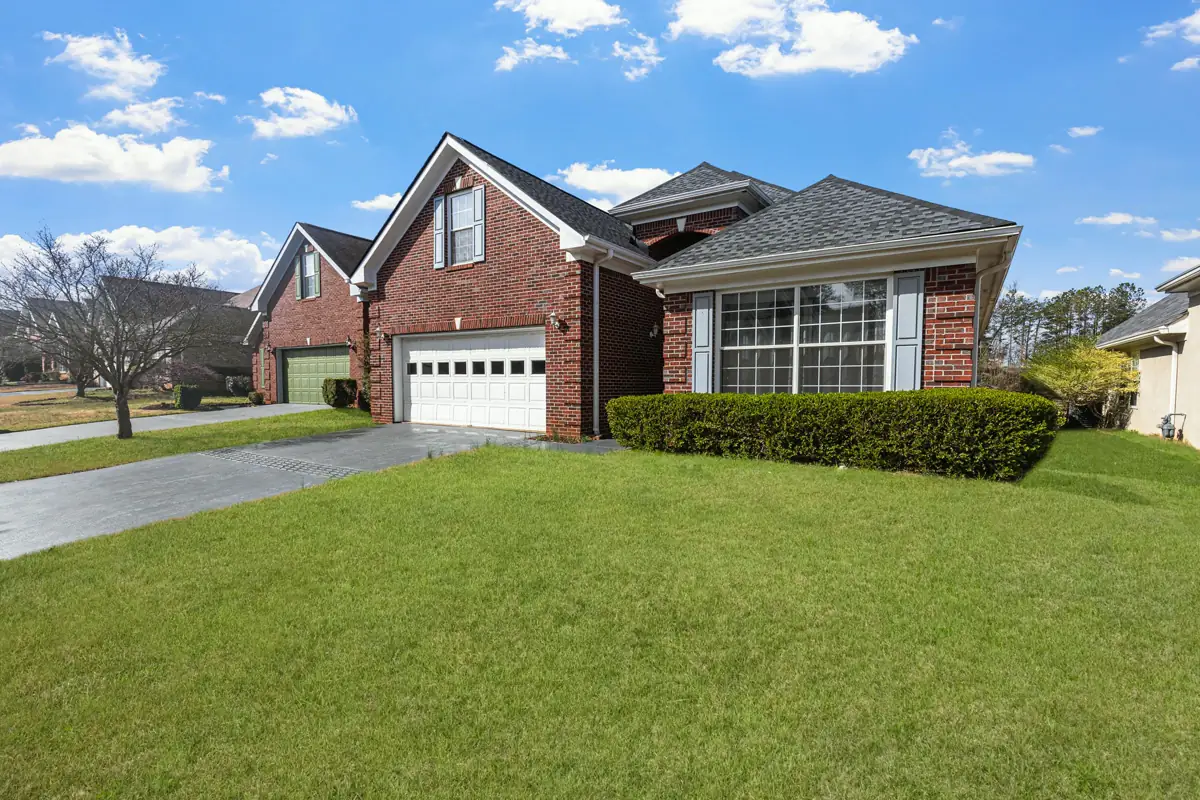 A red brick home with a front-facing garage gable, a wide picture window and a deep green front yard under a blue sky.