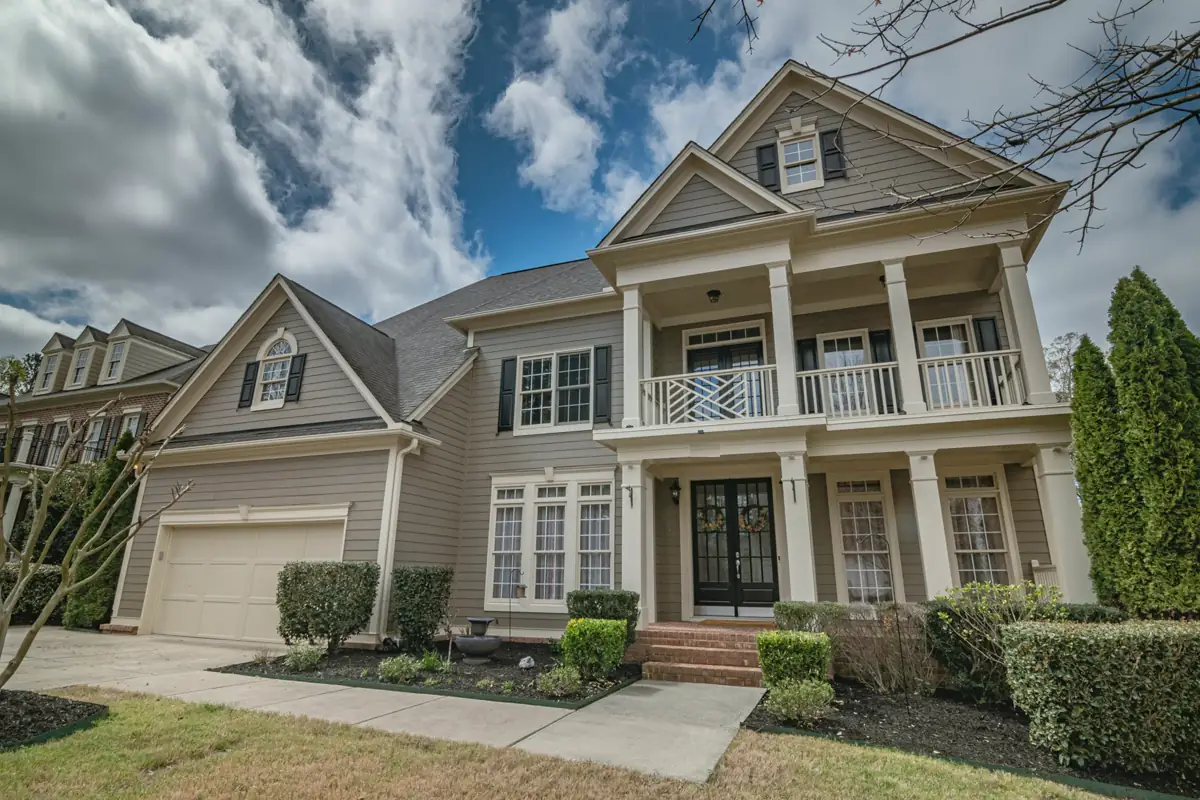 A taupe two-story home with a double front porch, cream columns, a balcony rail and an attached garage, framed by tall evergreens.